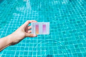 Hand holding a pool water testing kit over a swimming pool, with color charts for measuring pH and chlorine levels.