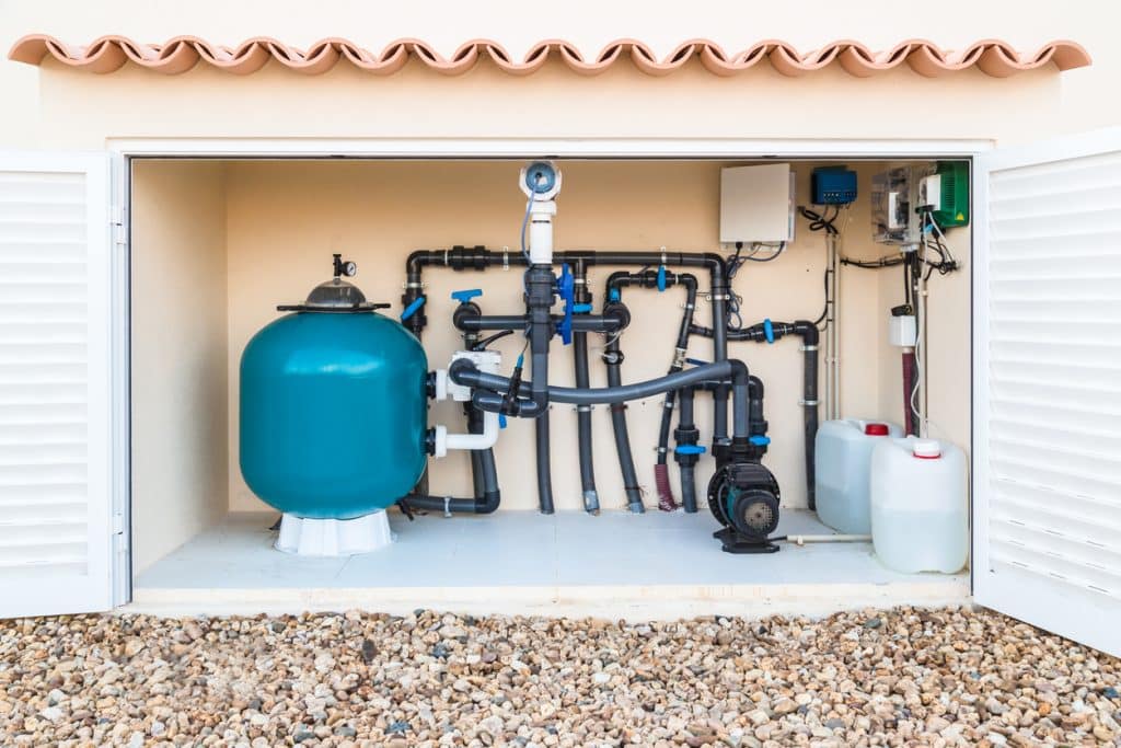 Swimming pool equipment system with a large filter tank, pump, connected pipes, valves, and chemical containers inside an outdoor equipment enclosure.